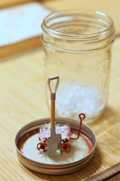 DIY Waterless Snow Globes with Kids Hot gluing a miniature shovel to the lid of a mason jar.
