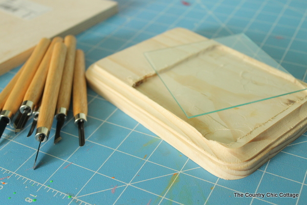 Plywood with recess cut and class tile on top, next to wood carving tools atop blue craft mat