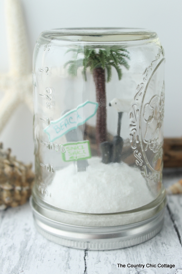 A beach sign inside a mason jar terrarium with a mini palm tree.