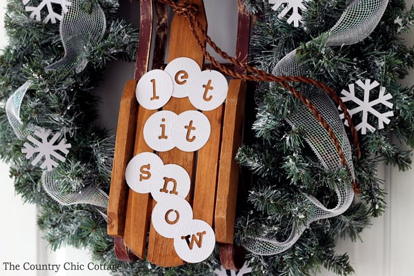 christmas wreath with sleigh attached in center and "let it snow" message on sleigh hanging on white door