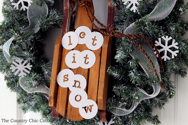 christmas wreath with sleigh attached in center and "let it snow" message on sleigh hanging on white door