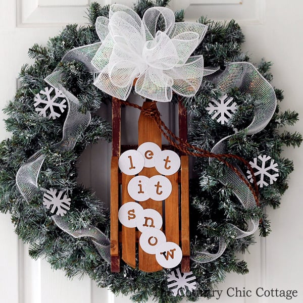 christmas wreath with sleigh attached in center and "let it snow" message on sleigh hanging on white door