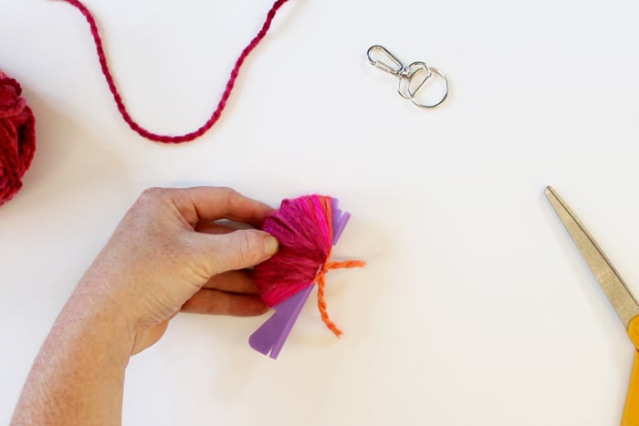 person wrapping pink yarn around small purple pom pom loom