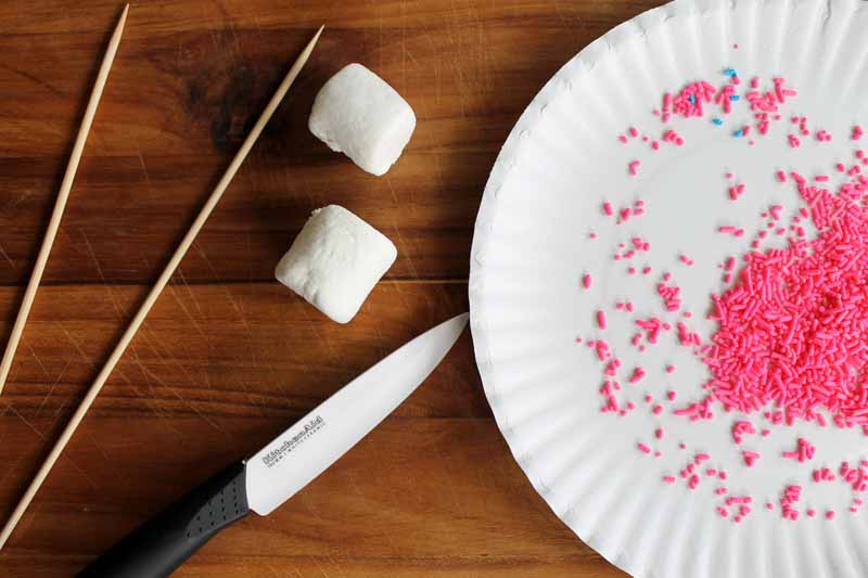 knife, marshmallows, wooden skewers next to a plate of pink sprinkles on wooden table