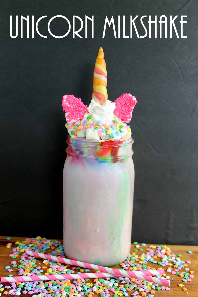 unicorn milkshake in a mason jar topped with whipped cream, sprinkles, candy ears, and a candy horn