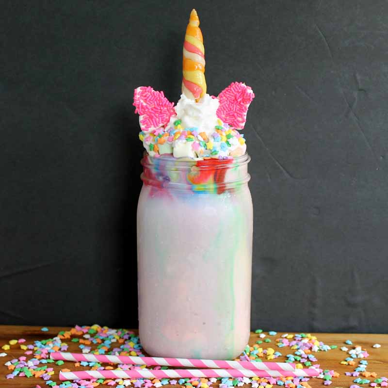 unicorn milkshake in a mason jar topped with whipped cream, sprinkles, candy ears, and a candy horn