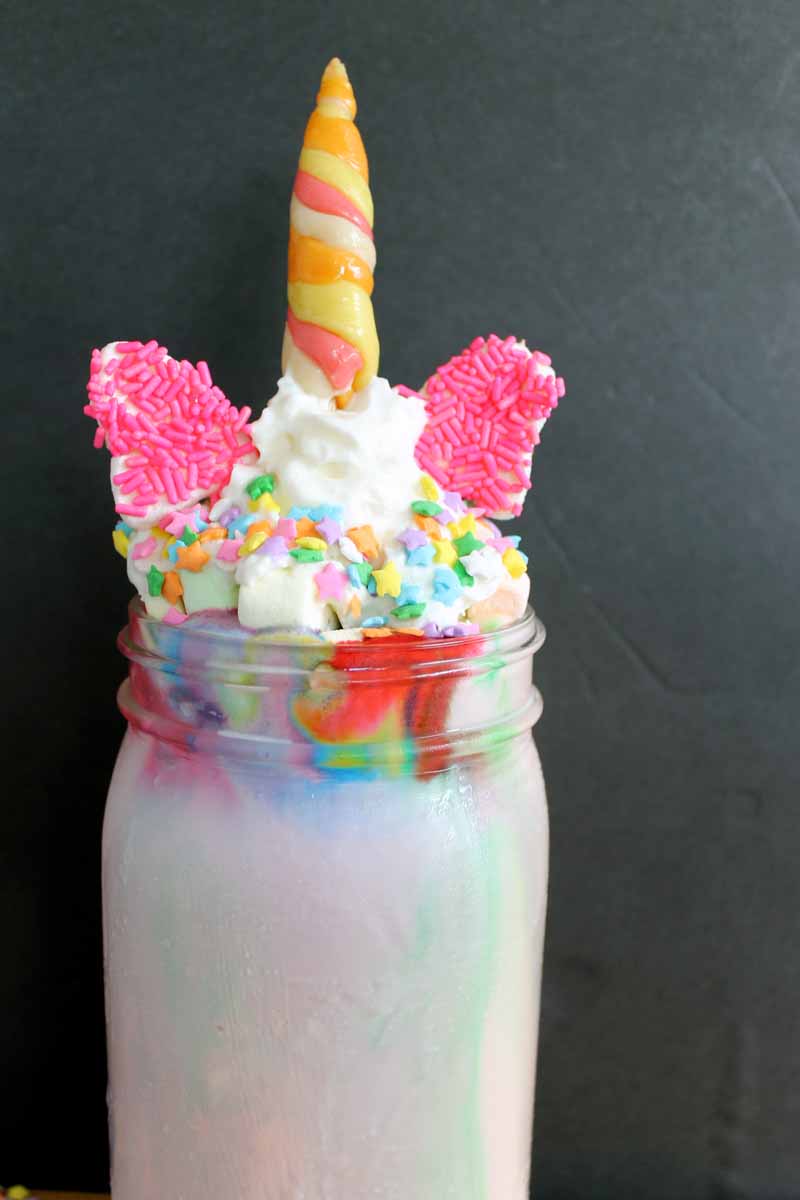 unicorn milkshake in a mason jar topped with whipped cream, sprinkles, candy ears, and a candy horn