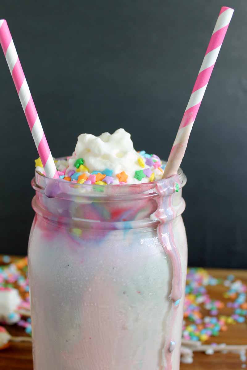 unicorn milkshake in a mason jar topped with whipped cream, sprinkles, candy ears, and a candy horn