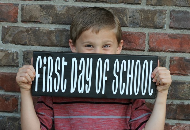 A young boy sitting in front of a wall holding first day of school black and white sign