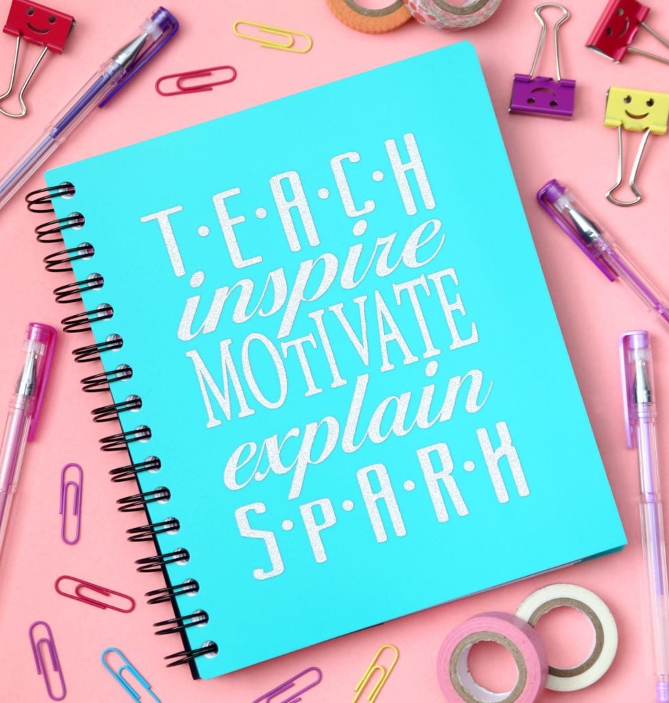 teach inspire motivate explain spark turquoise notebook on pink backdrop with school supplies scattered