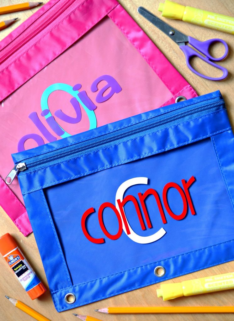 blue and pink pencil bags with vinyl monograms on a wood backdrop with purple scissors and glue sticks. 