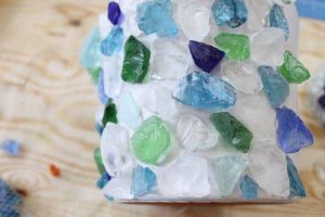 White pot decorated with blue, green, and clear sea glass on a table.