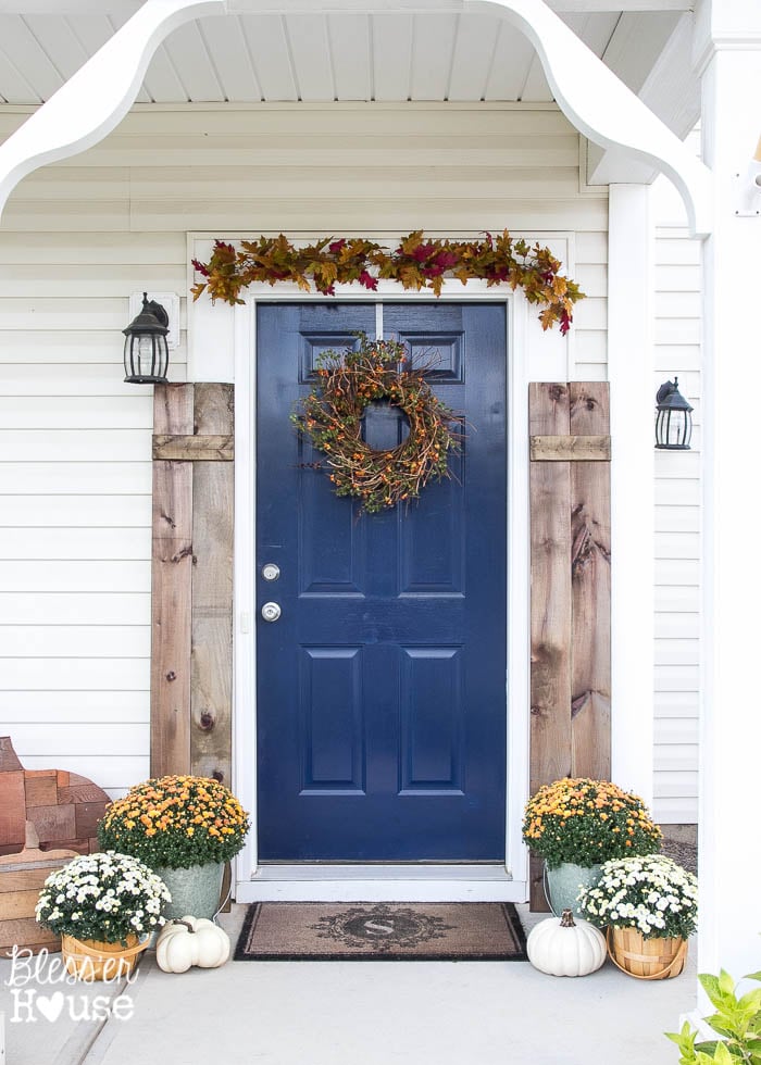 Front porch with blue front door, fall wreath, barnwood signs, mums and a leaf garland