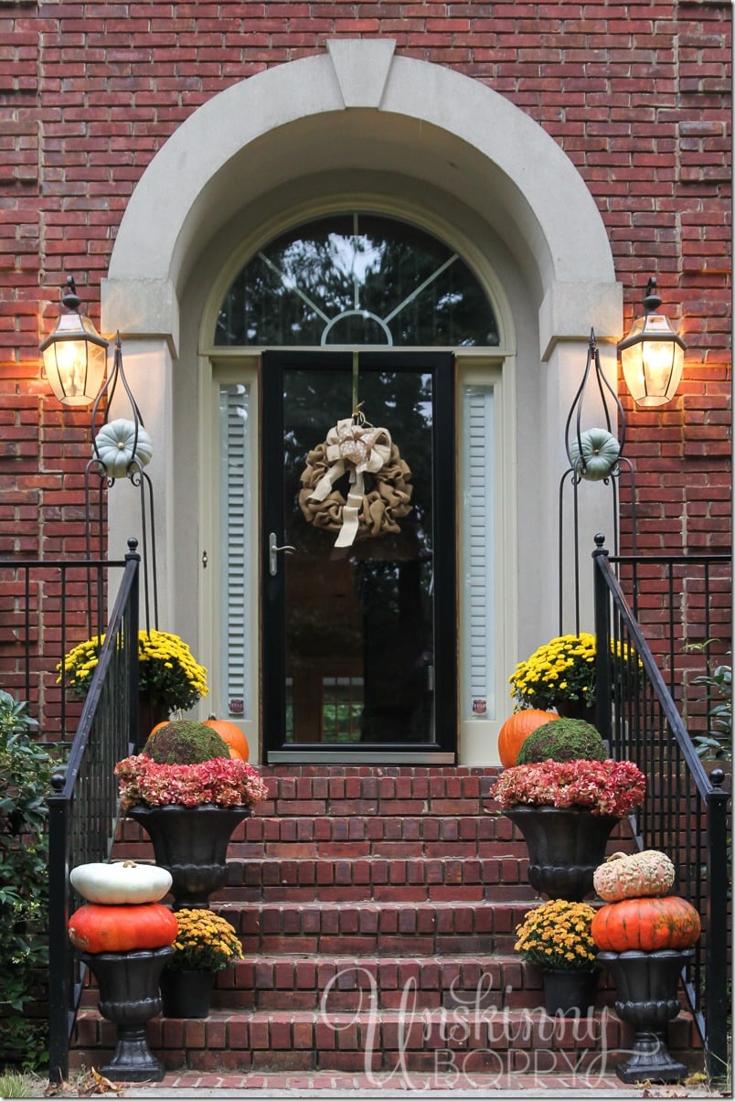 Front porch of brick house with black door. Fall wreath, potted mums, and heirloom pumpkins