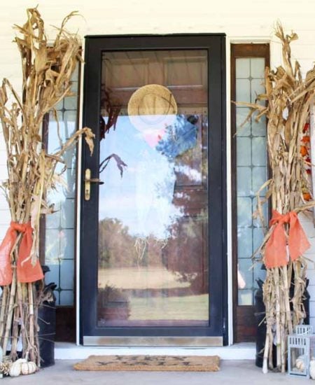 Farmhouse fall front porch with tied corn stalks on each side of a door and mini pumpkins