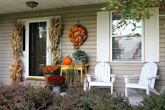 Fall front porch with fall wreath, corn stalks, yellow table with pumpkin and mums.